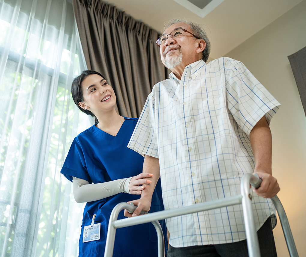 A nurse assists an elderly man using a walker in a brightly lit room.