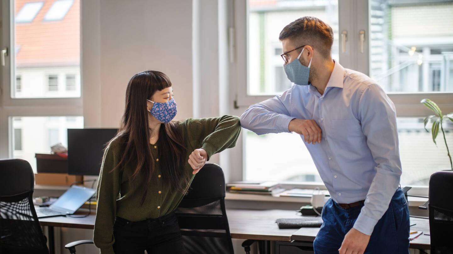 Two people in masks greet each other with elbow bumps in an office setting.