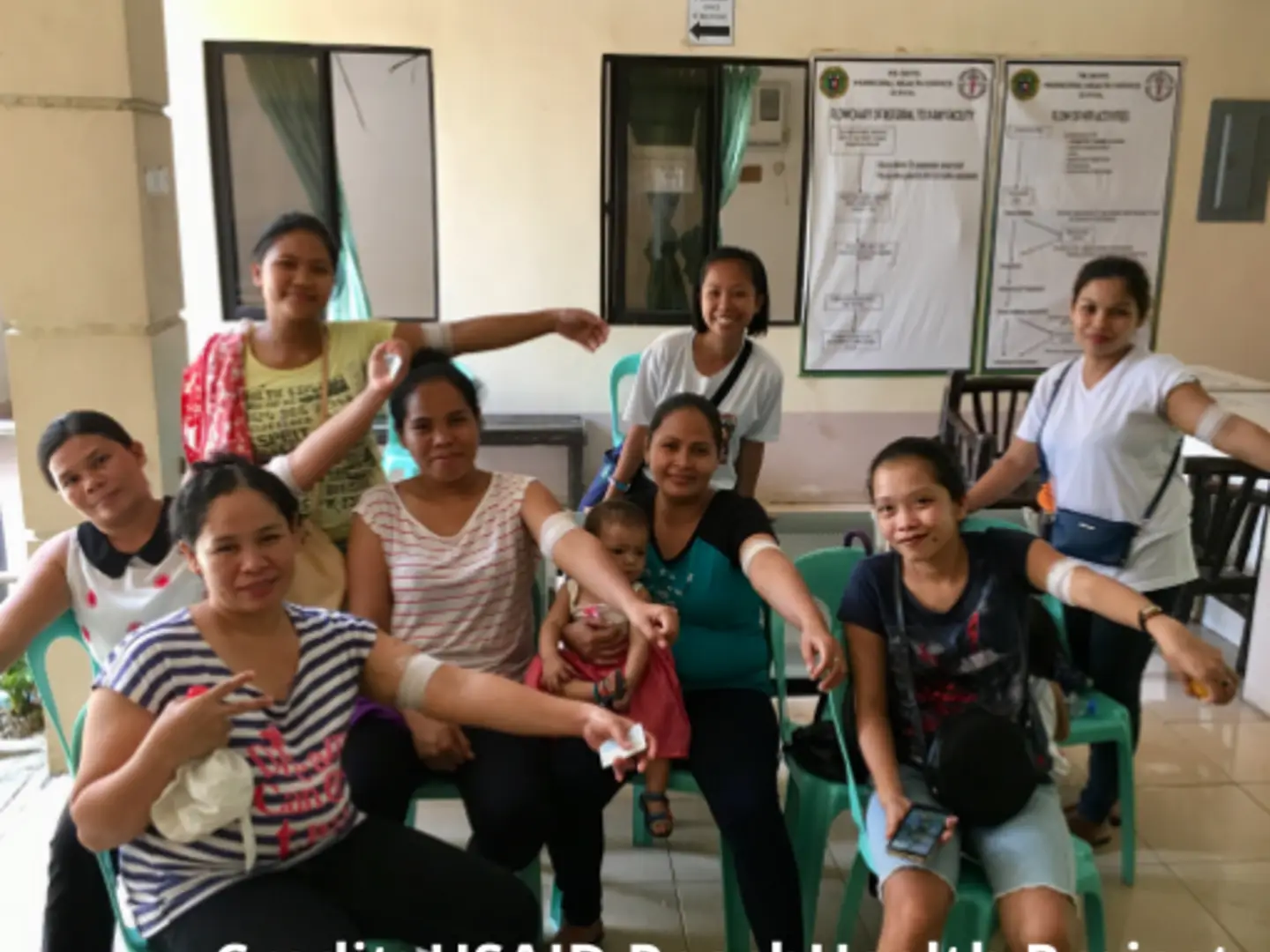 Group of women showing arms with bandages, sitting and smiling, indoors with informational posters on walls.