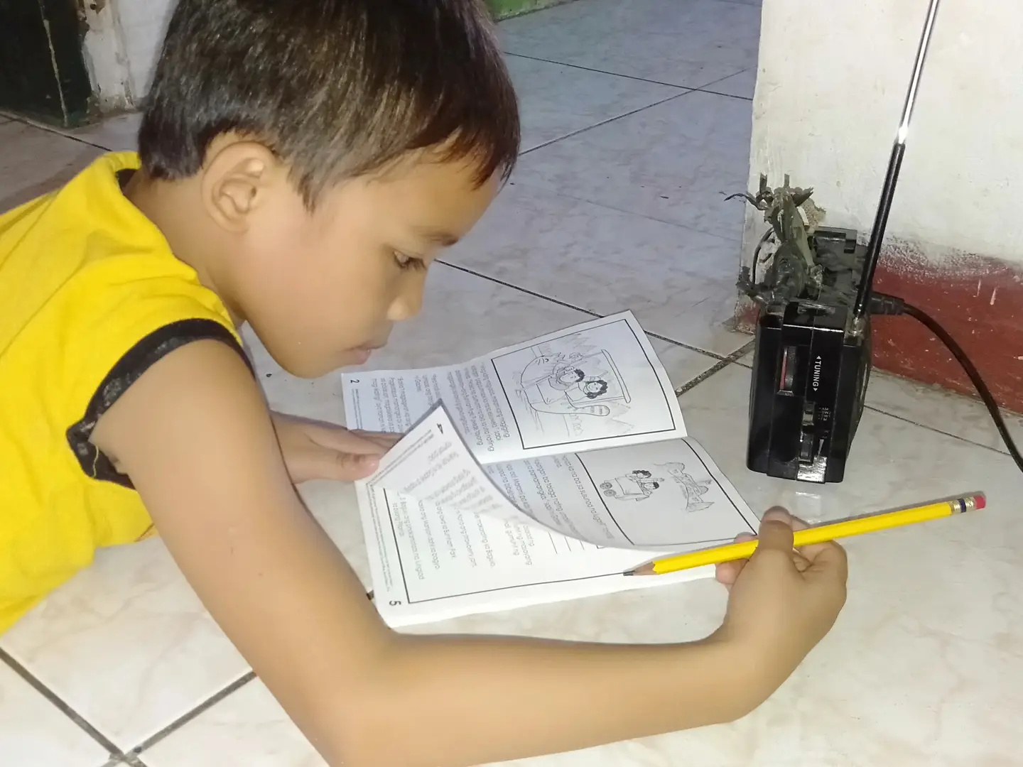 Child reading a book, holding a pencil on a tiled floor near a black radio.