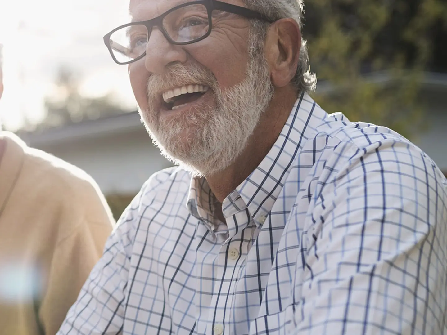 Smiling man wearing glasses and checkered shirt, sitting outdoors with blurred background of greenery.