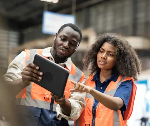 Two workers examine a tablet together in an industrial warehouse.