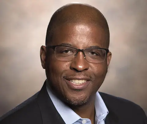 A smiling man with glasses poses in a professional setting, wearing a blazer over a shirt.