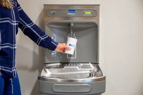 Person filling water bottle at wall-mounted water dispenser in public indoor setting.