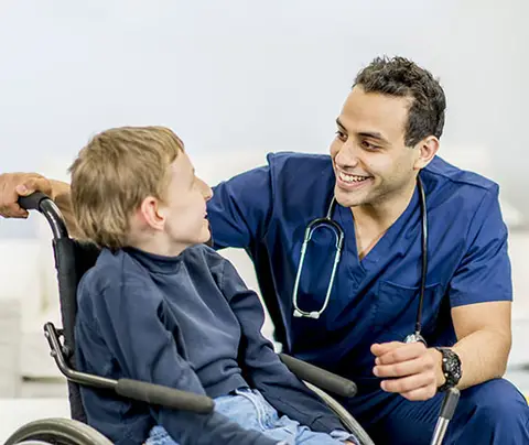 Healthcare professional smiling, comforting a child in a wheelchair in a brightly lit room.
