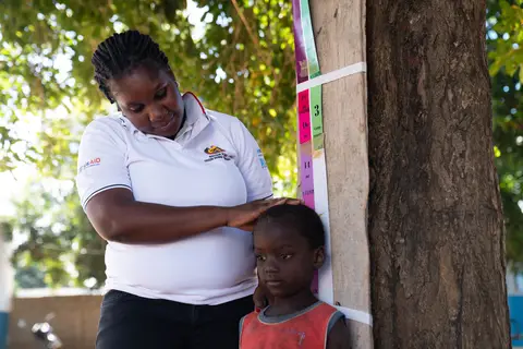 A woman measures a child's height against a tree in an outdoor setting.