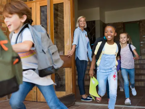 Children running outside a school entrance, carrying backpacks, with a smiling teacher holding the door open.