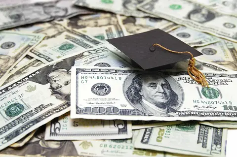 Graduation cap resting on assorted U.S. dollar bills, symbolizing education financing, on a money-covered surface.