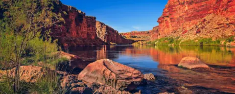 River flowing between steep, red canyon walls with greenery and rocks along the water's edge.