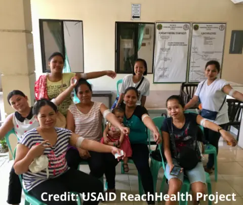Group of women showing arms with bandages, sitting and smiling, indoors with informational posters on walls.