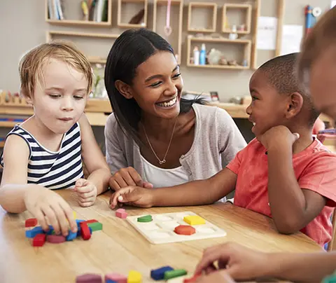 Teacher smiling, playing shape-sorting game with diverse children in a classroom setting.