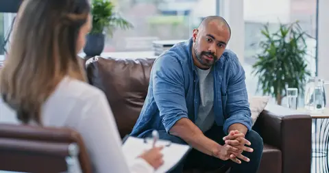 Man sitting on a couch talking to a woman with a notepad in an office setting.