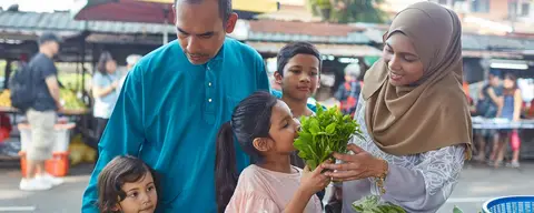 Family buying fresh vegetables at a market, surrounded by stalls and other shoppers.