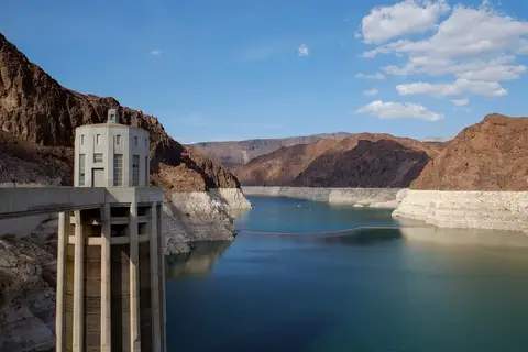 Dam structure spans a deep canyon filled with water; mountains and blue sky backdrop.