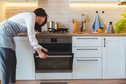 Woman opening oven door in modern kitchen with utensils and appliances on the countertop.