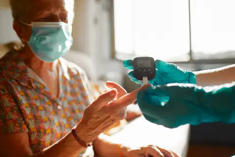 Elderly woman wearing mask gets blood sugar test by gloved hands in a sunlit room.
