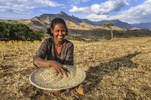 A woman sifting grains outdoors in a dry field with mountainous landscape in the background.