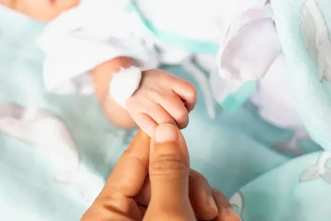 Baby's bandaged hand grasping an adult finger, lying on a light blue blanket.