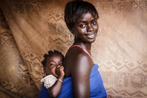 A woman carries a baby on her back, both smiling, against a patterned cloth background.