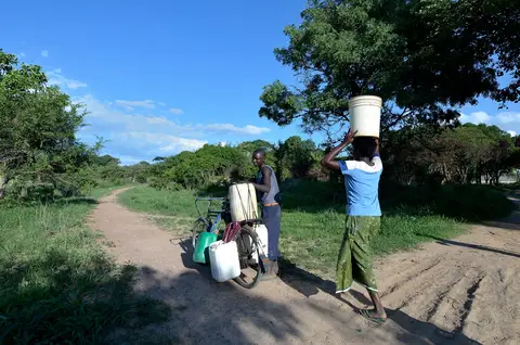 Two people transporting water containers, one on a bicycle, another balancing one on her head, along a dirt path surrounded by greenery.