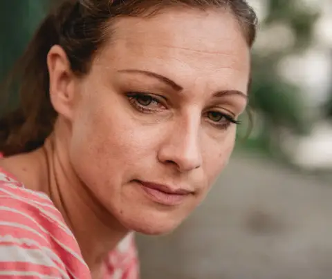 A woman wearing a red-striped shirt looking pensive, sitting outdoors with blurred greenery in the background.