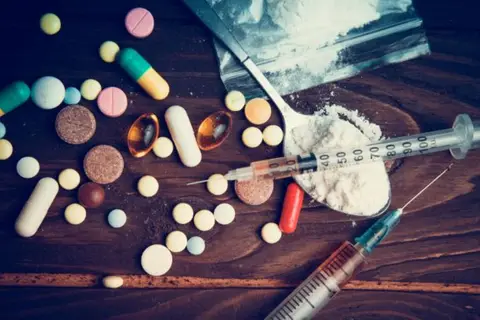 Various pills and syringes lie scattered on a wooden surface next to a bag of white powder.