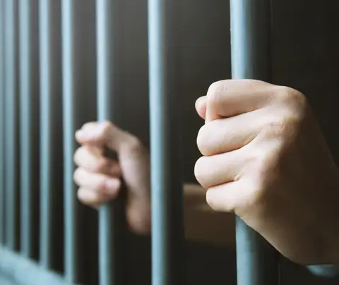 Hands gripping metal prison bars, inside a dimly lit cell.
