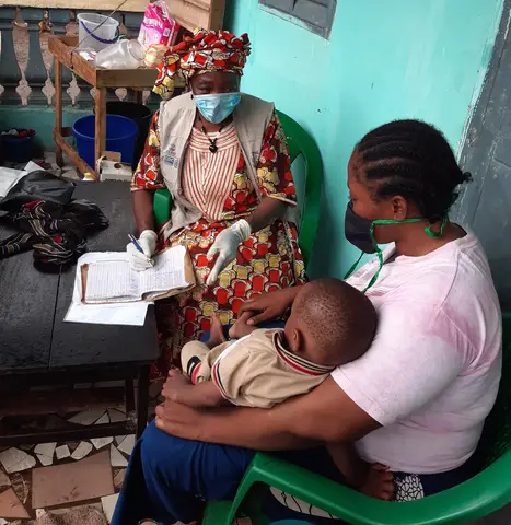 Healthcare worker records information while mother holds her child; both seated in a residential area.