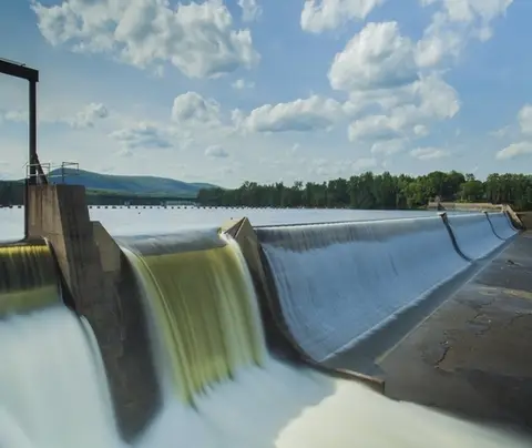 Water flows over a dam, surrounded by a lush forest and a cloudy sky.