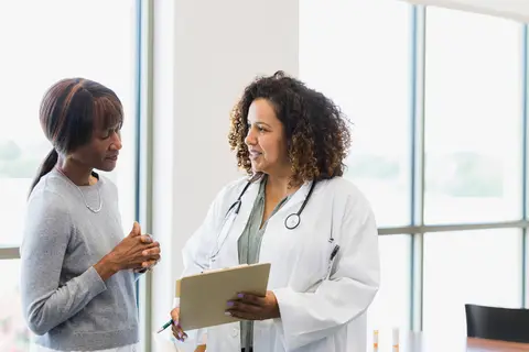 Doctor holding a clipboard, conversing with a patient in a bright, windowed medical office.