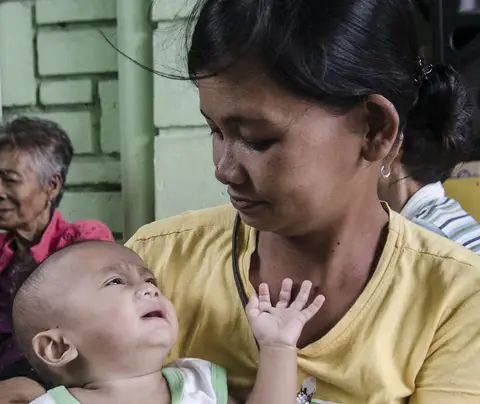 Baby reaching up to a woman who holds it, in a room with other people seated.