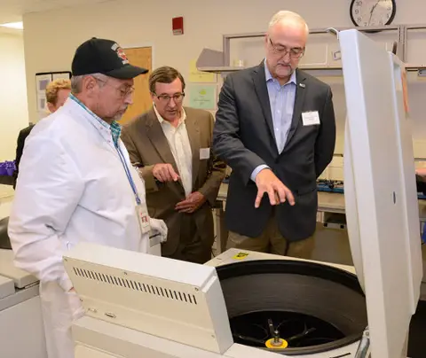 Three men examine an open centrifuge in a lab; one explains, others observe attentively.