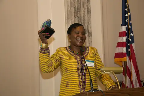 Smiling woman holding an award behind a podium beside an American flag in a formal setting.