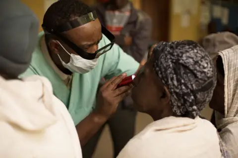 Healthcare worker checks patient's eyes with flashlight in a crowded indoor clinic.