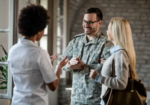 Soldier smiling, holding hands with a woman, talking to another woman in a medical uniform indoors.