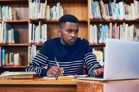 Man studying and writing notes at a desk in a library filled with books.