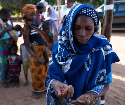 A woman in a blue patterned shawl examines pills; others queue in the background, outdoors.