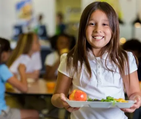 A girl smiles and holds a lunch tray in a school cafeteria with other children.