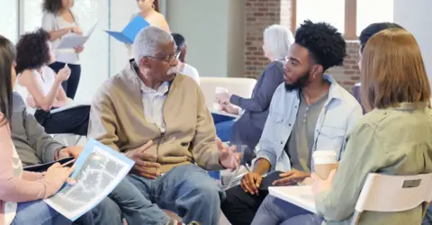 People having a discussion while holding documents in a spacious, well-lit room with other groups.