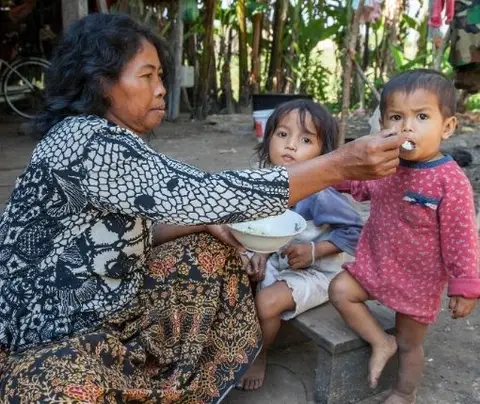 Woman feeding a young child with another child nearby, outdoors amidst trees and rustic surroundings.