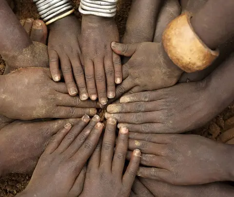 Hands forming a circle, suggesting unity, on a dirt ground; some wrists adorned with bracelets.