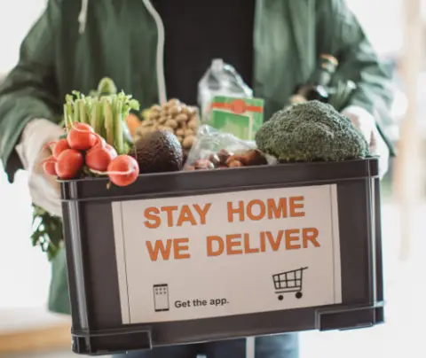 Person holding a crate of groceries with "STAY HOME WE DELIVER" printed on it.