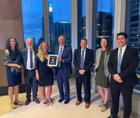 Group smiling, holding an award, in a modern office with cityscape background through large windows.
