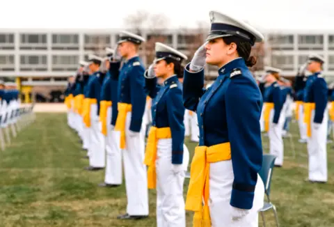 Cadets in blue uniforms and yellow sashes salute, standing in rows on a grassy field.