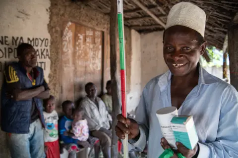 Man holding medical supplies and a colorfully striped rod; smiling in a rustic village setting.