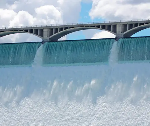 Water cascades over a dam, above which an arched bridge spans against a cloudy sky backdrop.