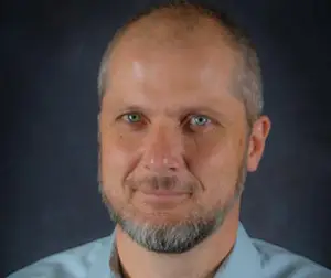 A bald man with a short beard smiles slightly against a dark, solid background.
