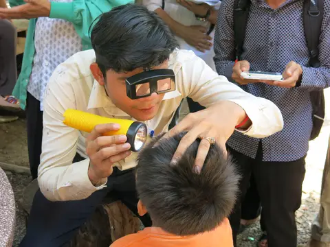 Man inspects child's scalp with flashlight and loupe, surrounded by onlookers outdoors.