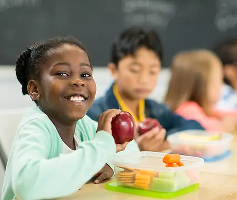 A smiling girl holds an apple, enjoying lunch with classmates in a classroom setting.