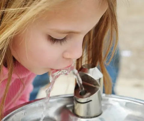 A girl drinks from a metal water fountain in a public outdoor area.